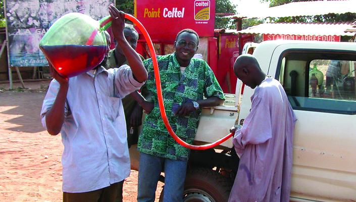 3 men in Chad fill their white pickup's gas tank by siphoning fuel from a glass bottle