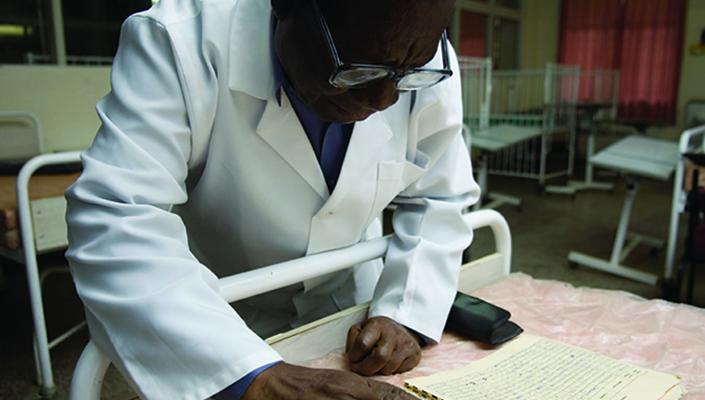 Clinician Redson Makasa, wearing a lab coat, reviews a paper ledger of measles cases