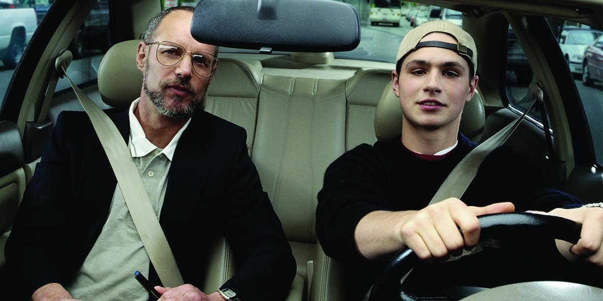 a teen wearing a backward baseball cap sits behind the steering wheel; his father sits in the passenger seat.