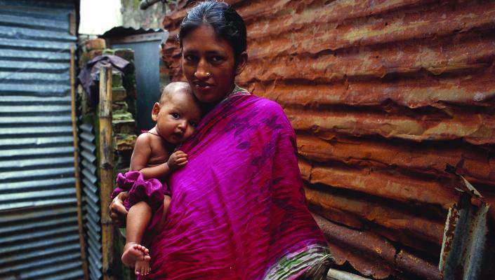  mother in Bangladesh wearing a magenta scarf holds her infant on her hip