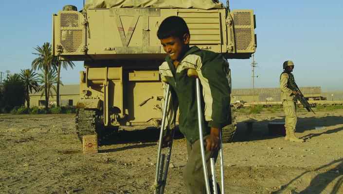 A young teen on crutches walks by a military vehicle guarded by a soldier