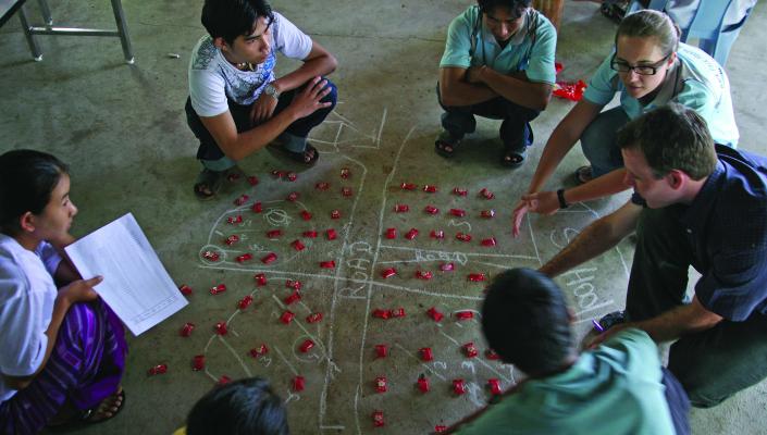 Luke Mullany and Kate Teela squat on the floor as they teach interval sampling to Burmese surveyors, using red candy packets on a chalked diagram