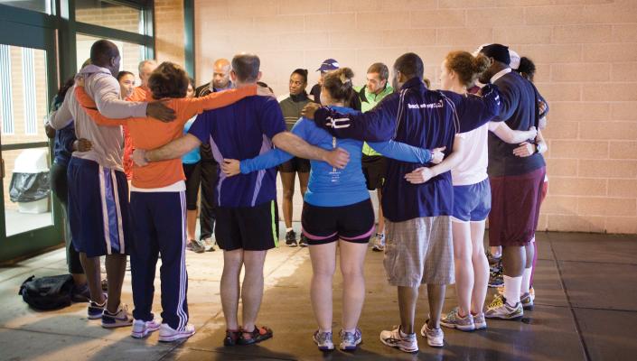 a circle of about 15 people in athletic gear stand in a circle with their arms around each other