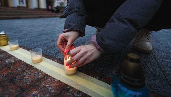 a person kneels to light a candle in a row of candle holders lined up on the street.