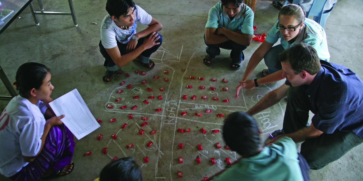 Luke Mullany and Kate Teela squat on the floor as they teach interval sampling to Burmese surveyors, using red candy packets on a chalked diagram