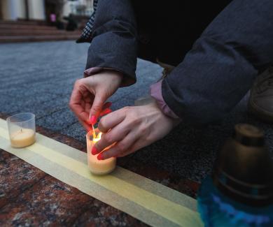 a person kneels to light a candle in a row of candle holders lined up on the street.